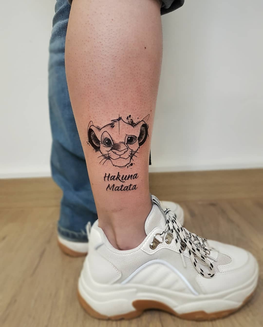 The tattoo features a cartoon lion cub Simba alongside the text "Hakuna Matata" on a person's leg